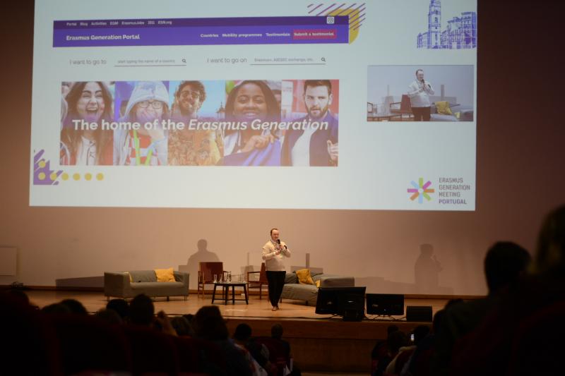 A picture of a person on a stage, speaking into a microphone. Behind them, projected on a big screen, is the front page of the newly launched Erasmus Generation Portal.