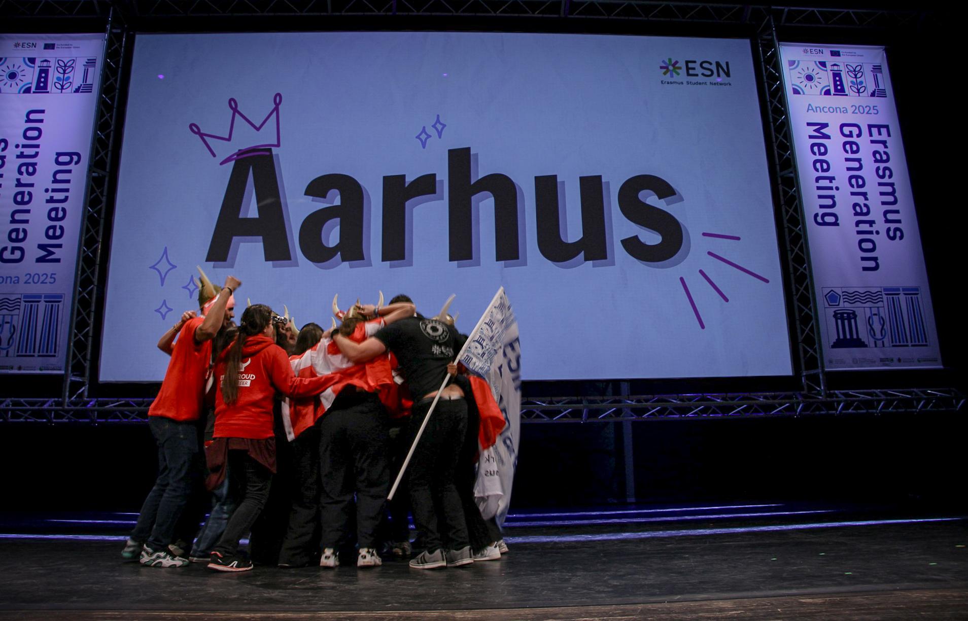 A group of people hugging and celebrating on a stage in front of a big screen with "Aarhus" on it.