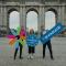 Three people, standing in front of a large stone arch monument, each holding oversized colourful props: the Erasmus Student Network logo, a circular Ryanair–ESN logo, and a rectangular Ryanair sign.
