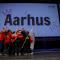 A group of people hugging and celebrating on a stage in front of a big screen with "Aarhus" on it.