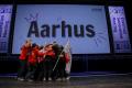 A group of people hugging and celebrating on a stage in front of a big screen with "Aarhus" on it.