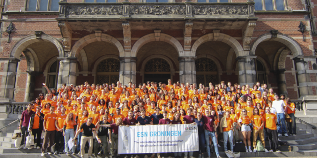 A big group of people in orange shirts posing for a picture and holding a flag of ESN Groningen.