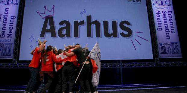 A group of people hugging and celebrating on a stage in front of a big screen with "Aarhus" on it.